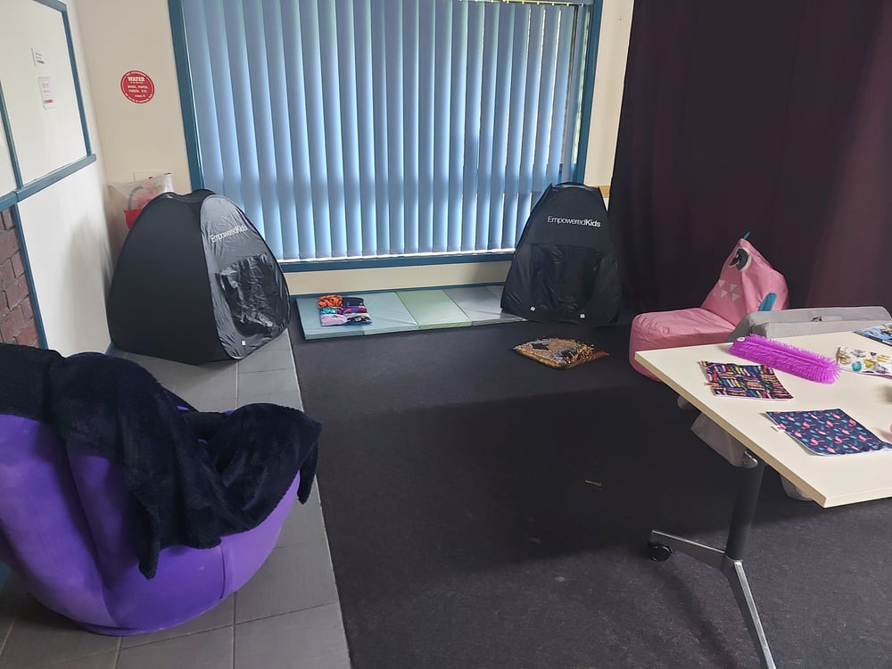 A small room with a bean bag, a comfortable chair and a small black tent. The room is lit with filtered natural light and has a table of fidget items in the corner.
